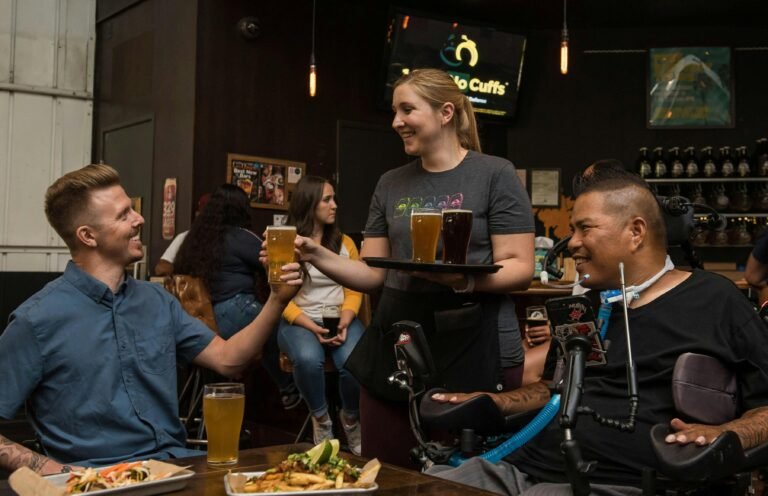 A waitress serves drinks to friends, including one in a wheelchair, in a lively bar setting.